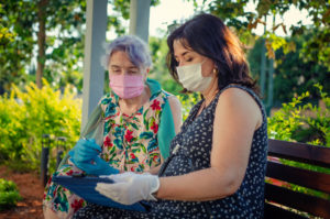 senior woman and middle-age woman sitting on a park bench wearing masks and gloves while filling out paperwork on clipboard
